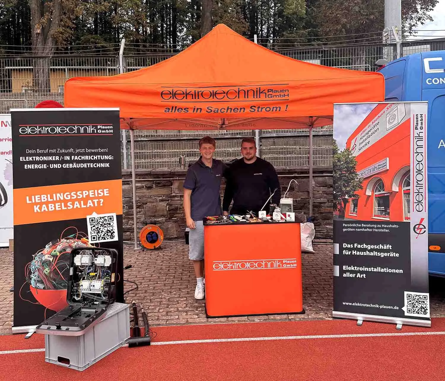Messestand mit oranger Plane elektrotechnik Plauen GmbH, zwei Mitarbeiter hinter der Theke, daneben Rollups zur Ausbildung