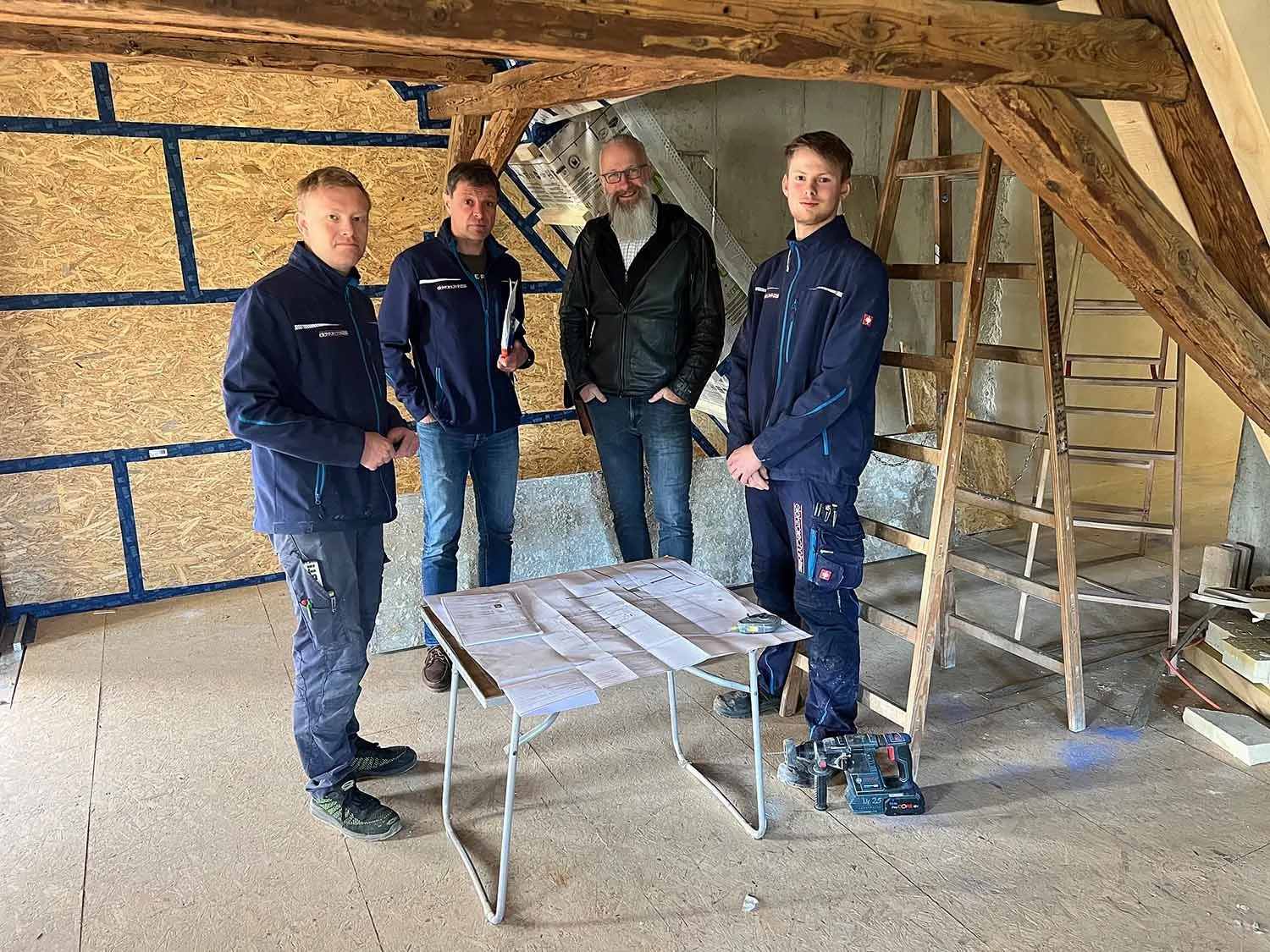 Vier Männer im Dachgeschoss unter Holzbalken an einem Tisch mit Plänen