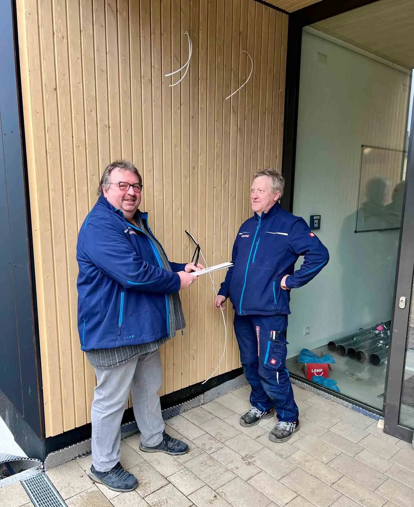 Zwei Mitarbeiter in Firmenjacken vor einer Holzfassade, aus der Wand ragen Kabel